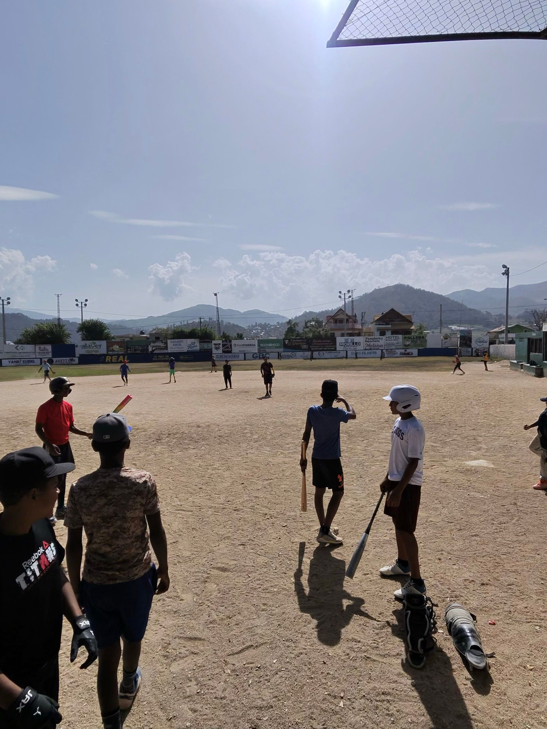 Kids with bats and one helmet on the dirt infield in Constanza, mountains behind the outfield wall
